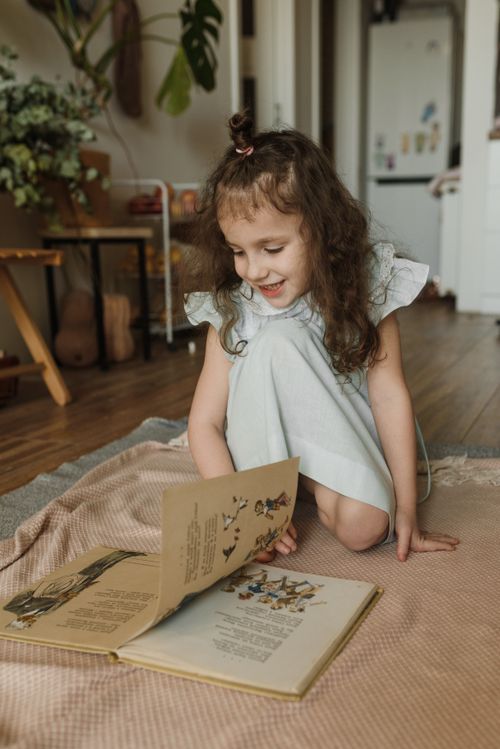Free Charming young girl reading a book indoors, sitting on a soft rug with a joyful smile. Stock Photo