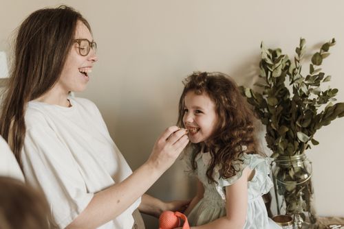 Free A mother joyfully feeds her smiling daughter a cookie inside their cozy home. Stock Photo