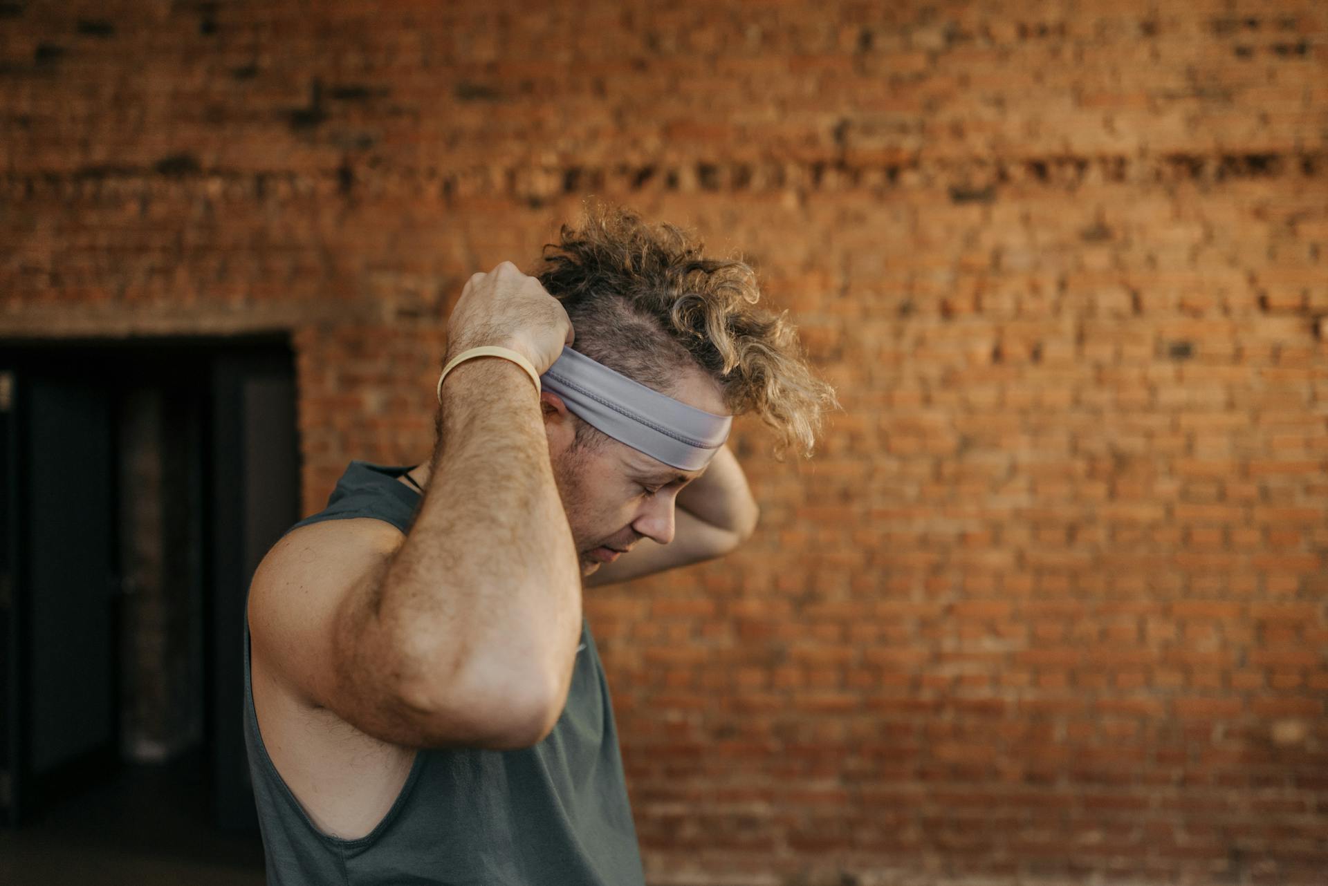 A man wearing a tank top adjusts his headband indoors against a brick wall.