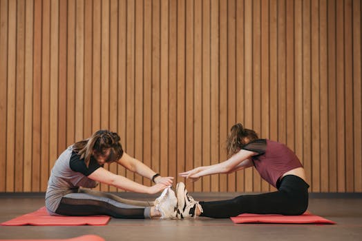 Two women performing seated stretches together on yoga mats indoors.