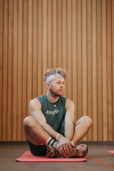 A man sits cross-legged on a yoga mat in a gym, wearing activewear and a headband.