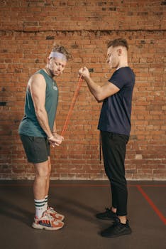 Two men exercising with resistance bands against a brick wall background.