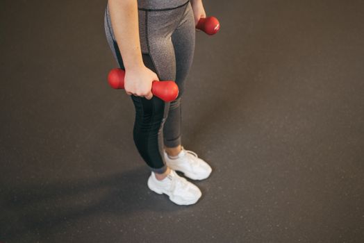 Female fitness enthusiast standing with red dumbbells on a gym floor.