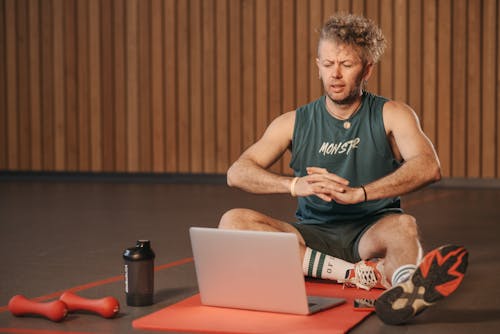 Free Man Sitting on Exercise Mat While Looking at the Screen of a Laptop Stock Photo