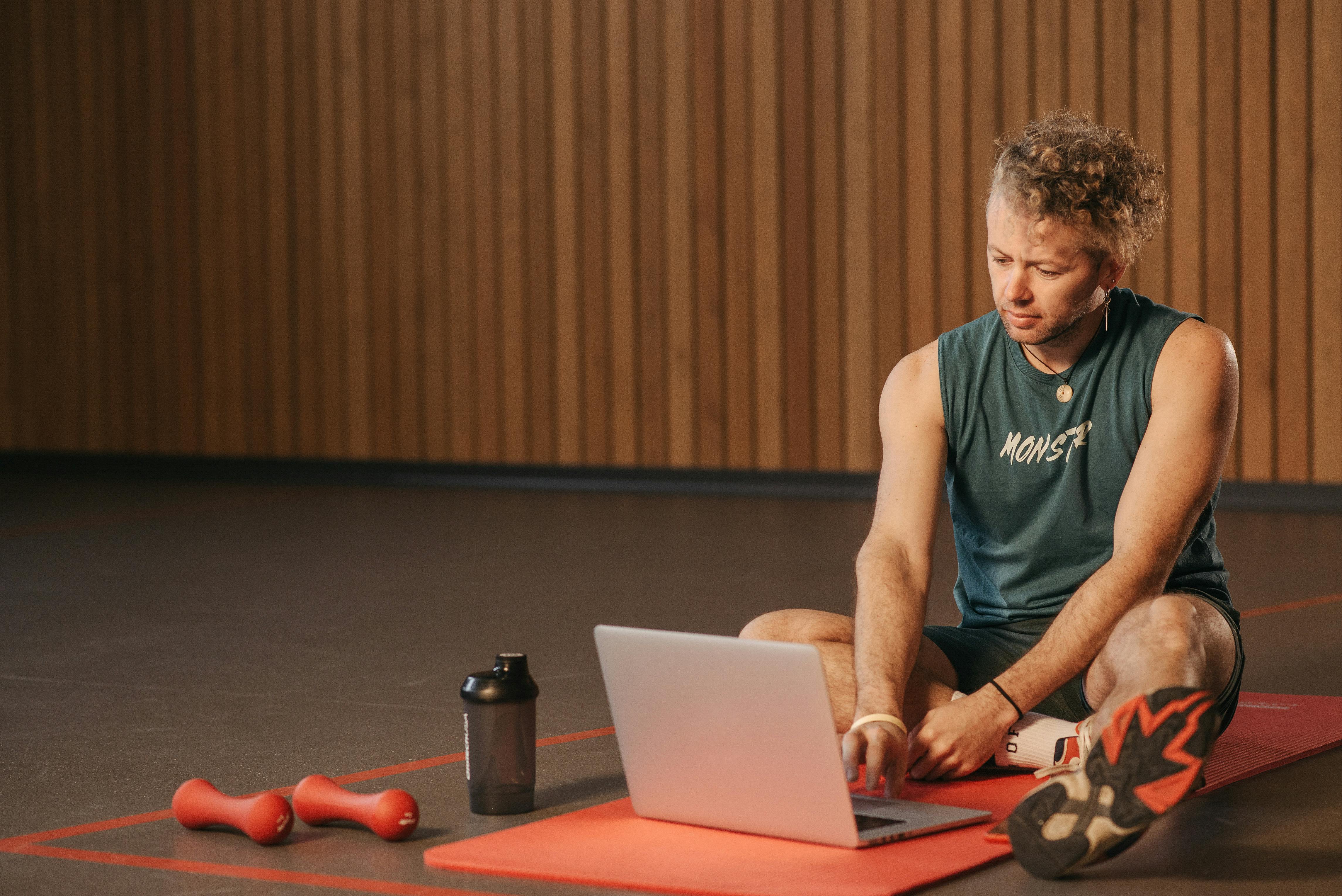 Man sitting on exercise mat with laptop, surrounded by dumbbells, creates a unique fitness workspace.
