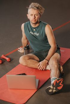 Man in gym attire seated on a yoga mat using a laptop, exemplifying multitasking in fitness.