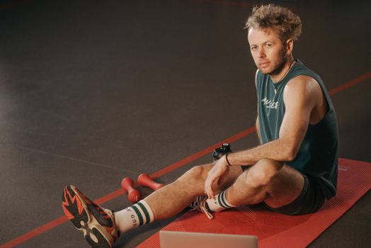 Adult male in sportswear sitting on a mat in a gym setting with dumbbells nearby.