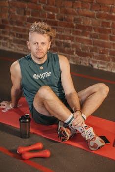 Man resting on a red exercise mat with dumbbells in a brick-walled gym.
