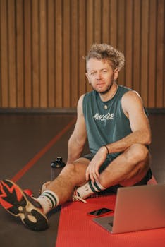 Adult man with facial hair sits on a yoga mat with a laptop, exercising indoors.