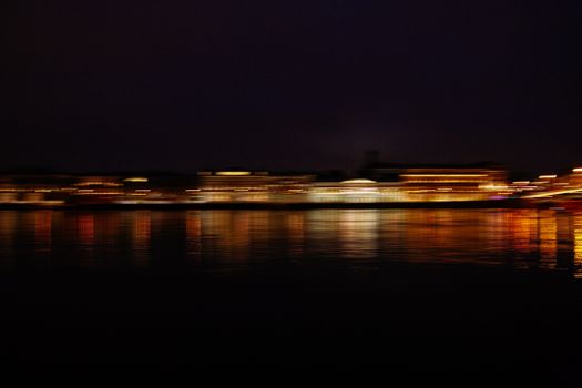 A vibrant, blurred cityscape with illuminated reflections over water at night.