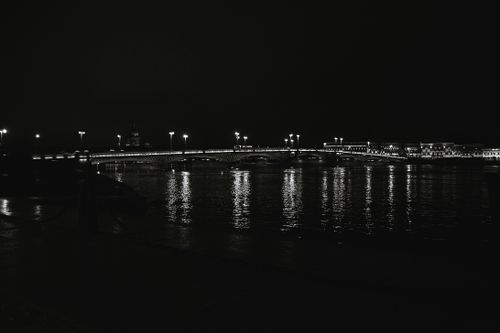 Free A serene black and white nighttime view of a lit bridge reflecting on a calm river. Stock Photo