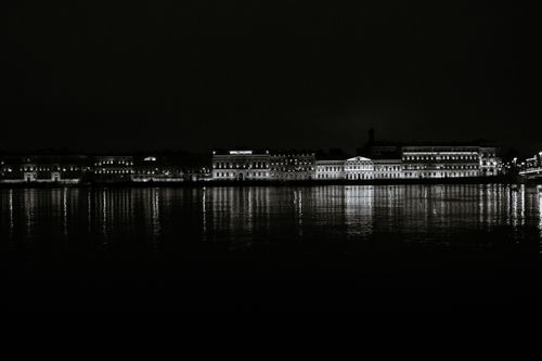 Free Monochrome cityscape featuring illuminated waterfront buildings reflecting on a calm river at night. Stock Photo