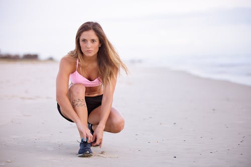 Free Woman in Pink Top and Black Shorts Lacing Her Shoes on Sea Shore Stock Photo