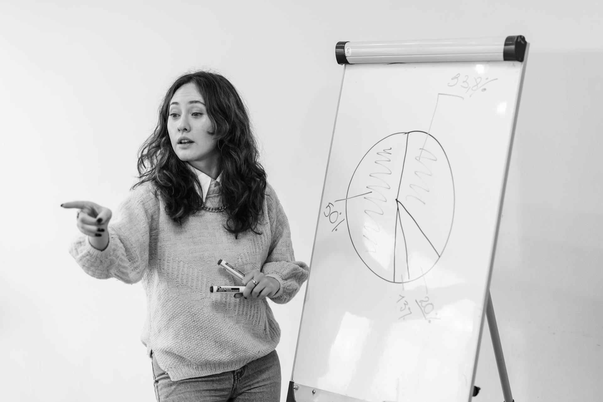 Grayscale Photography of a Woman Talking while Standing Beside the White Board