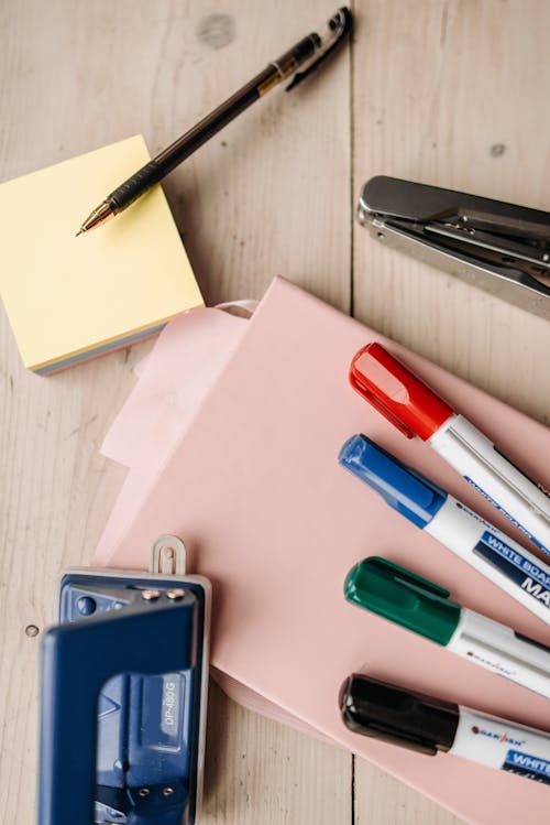 Colored Markers Lying on a Notebook next to Sticky Notes, Pen and a Stapler