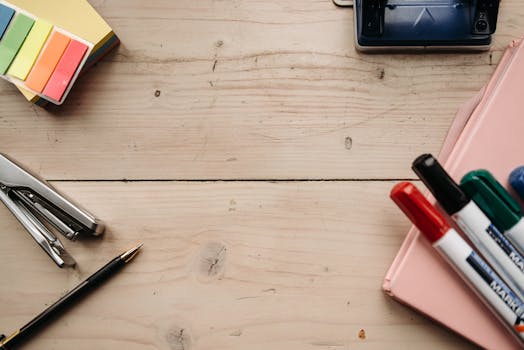 Top view of office supplies including markers, stapler, and notepad on a rustic wooden table.
