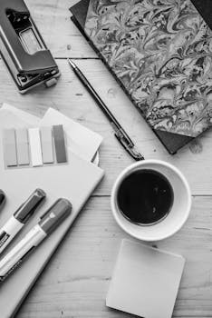 Monochrome flat lay of office supplies including coffee cup, notebook, and pens on a wooden desk.