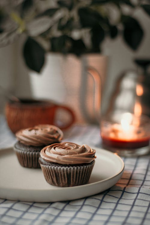 Delicious baked cupcakes in paper cups on plate placed near burning candle kettle against houseplant