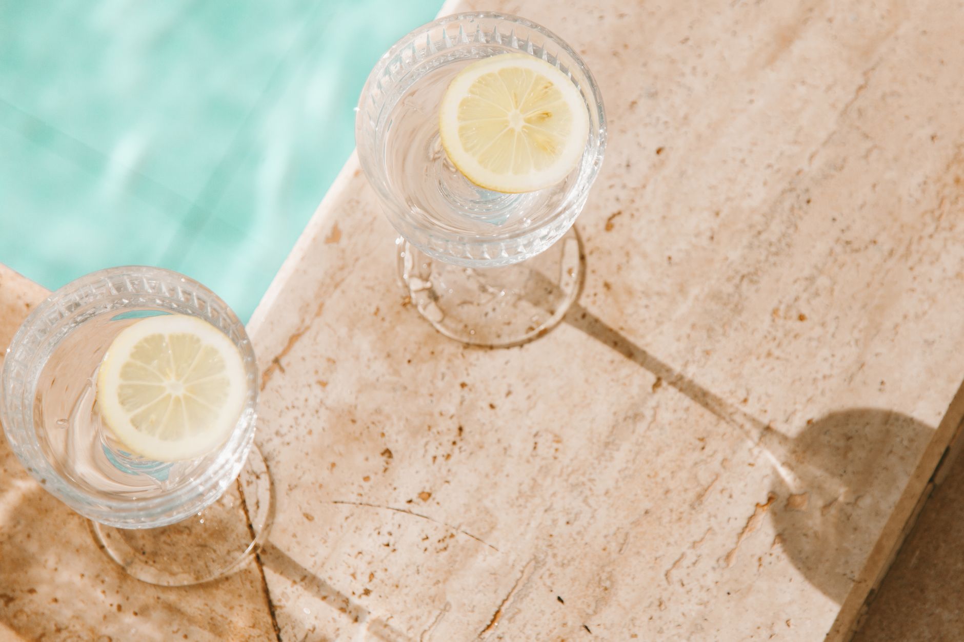 Two lemon water glasses on a sunlit concrete surface by a pool, perfect for summer vibes.