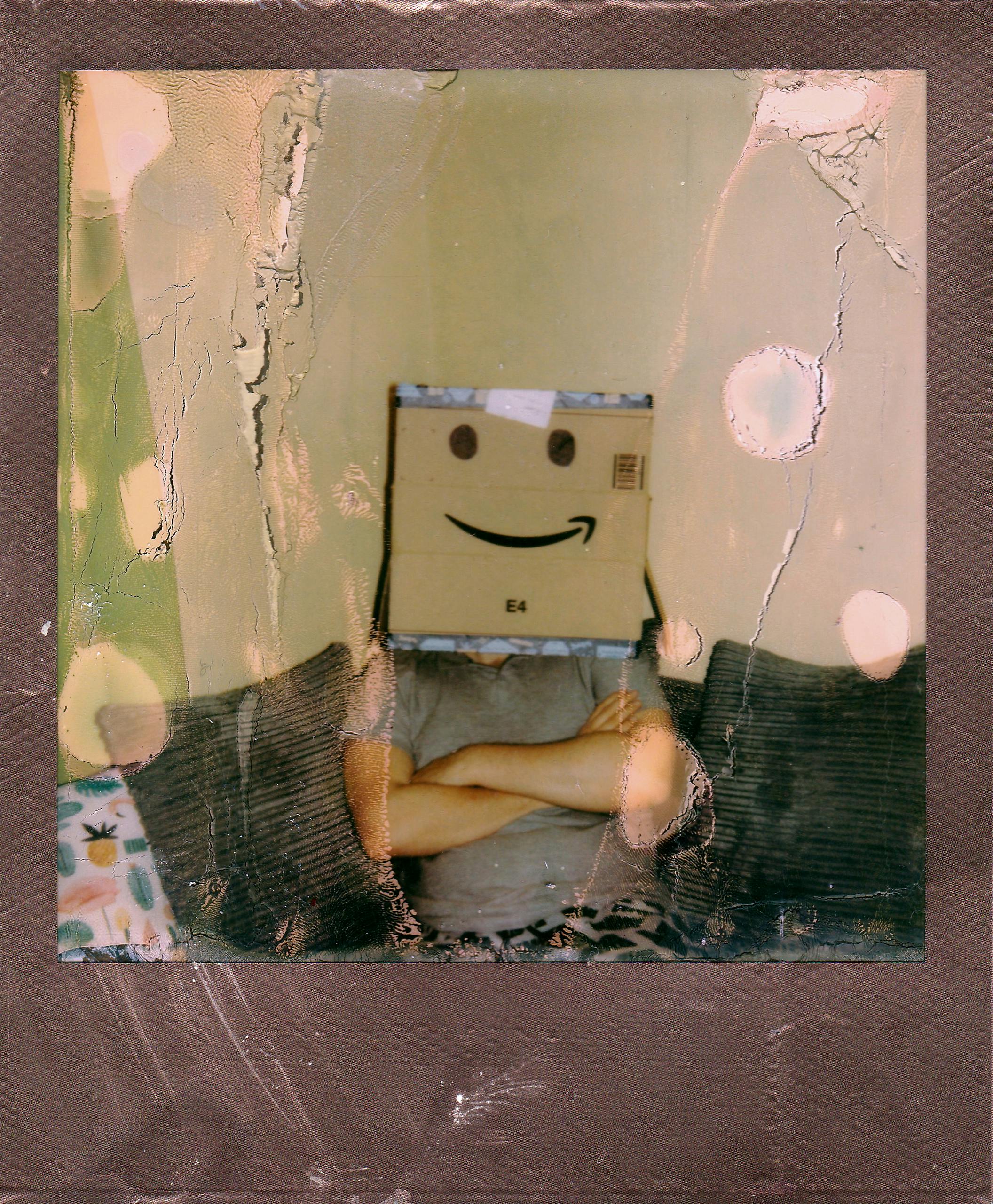 Artistic portrait of a person with a cardboard box head featuring a smiley face, arms crossed in a vintage style.