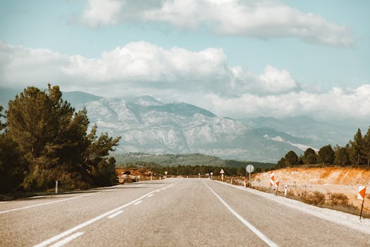 A serene highway leading through lush trees and majestic mountains under a cloudy sky.