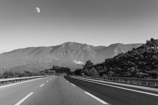 Black and white image of an empty highway with mountains and moon in the background.