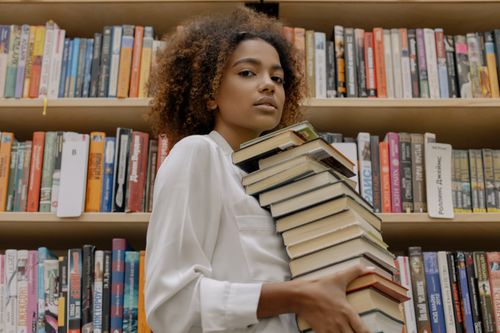 Free Confident young woman holds stack of books in a library setting. Stock Photo