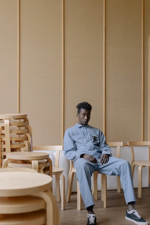 Man in a Blue Jumpsuit Sitting in a Room Among Chairs and Stools