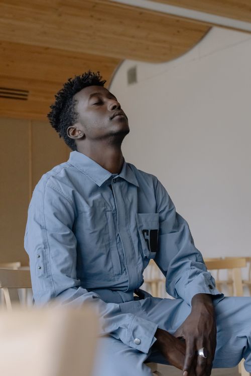 Free African American man in blue outfit sitting with eyes closed, looking relaxed in a modern room. Stock Photo