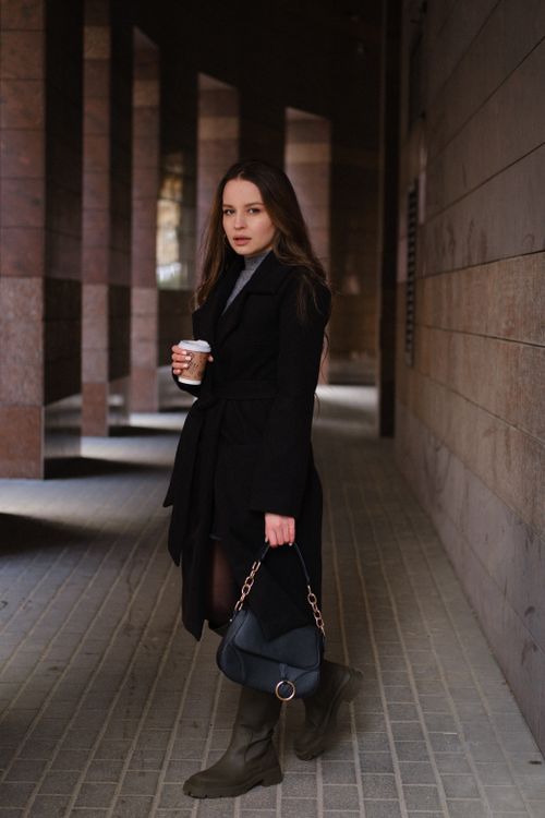 Free Full length of serious young female in stylish coat with purse and paper cup of coffee standing on town street near building with columns and looking at camera Stock Photo