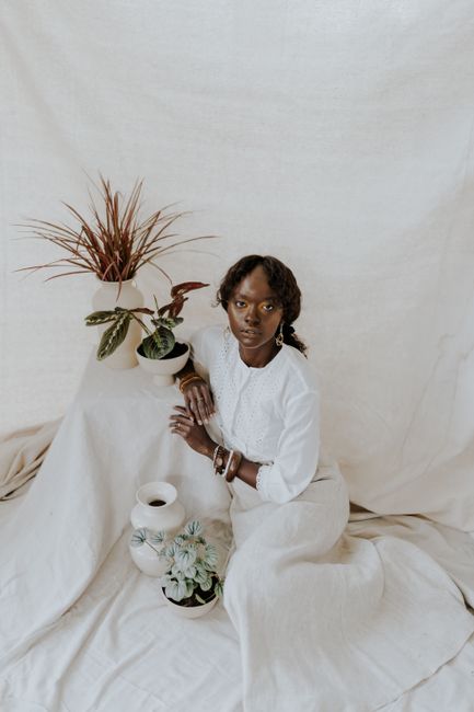 From above of content African American female in long white dress looking at camera on white fabric near potted plants