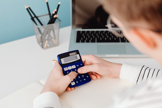 Close-up of hands using a blue calculator at a desk with a laptop and stationery.