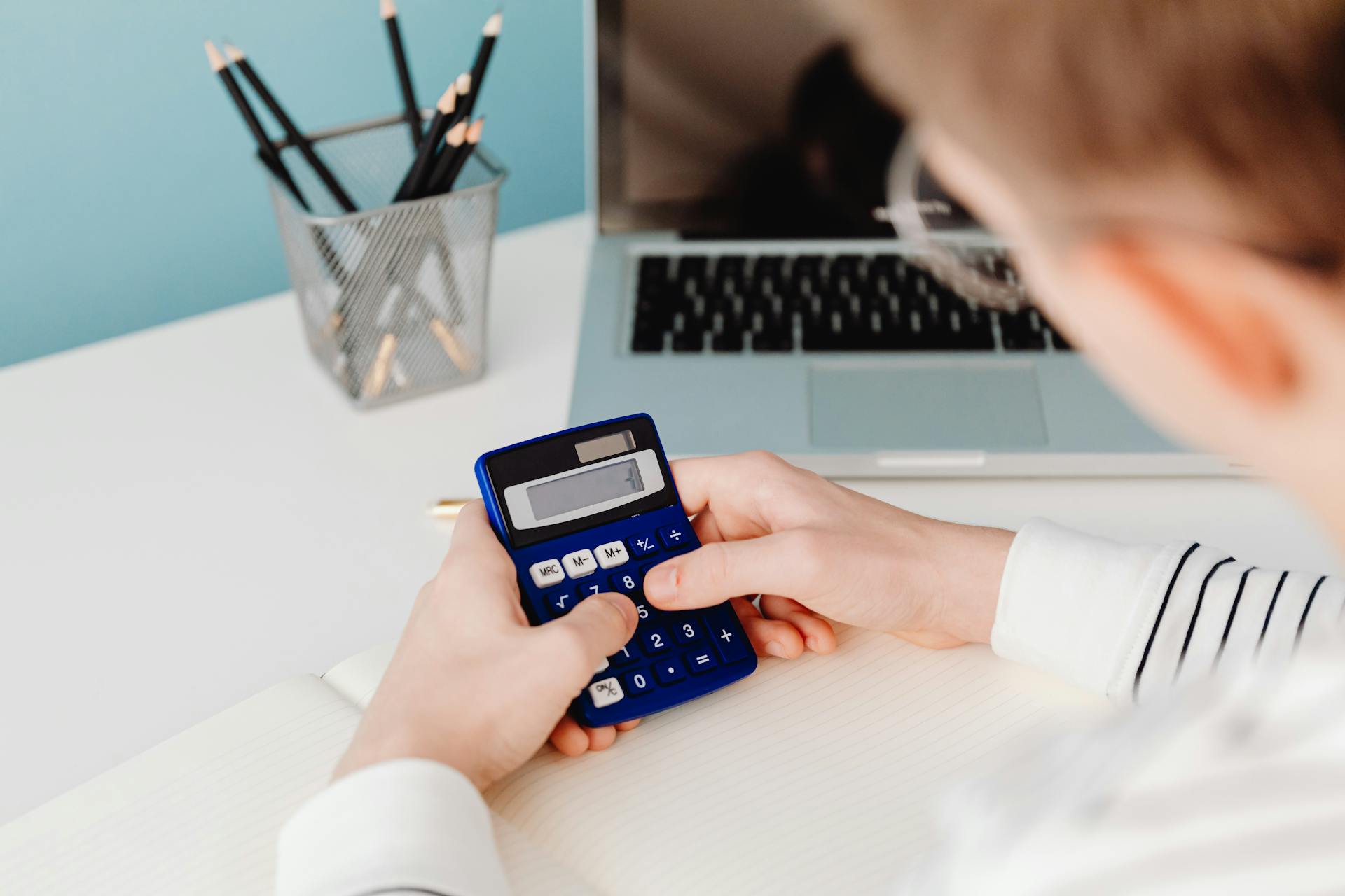 Close-up of hands using a blue calculator at a desk with a laptop and stationery.