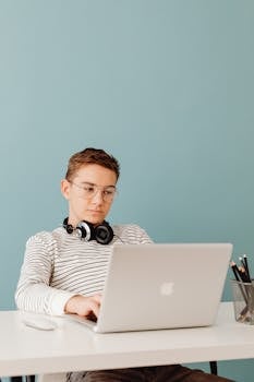 A young man using a laptop with headphones around his neck, sitting against a blue background.