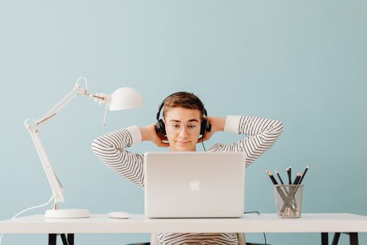 A young man with headphones and eyeglasses relaxes at a desk with a laptop in a modern indoor setting.