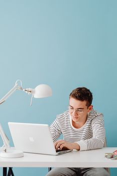 Young adult focused on his laptop at a modern home office with blue wall.
