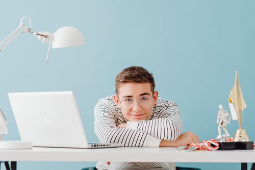Free Young man with glasses leaning on a desk with a laptop and trophies, against a teal background. Stock Photo