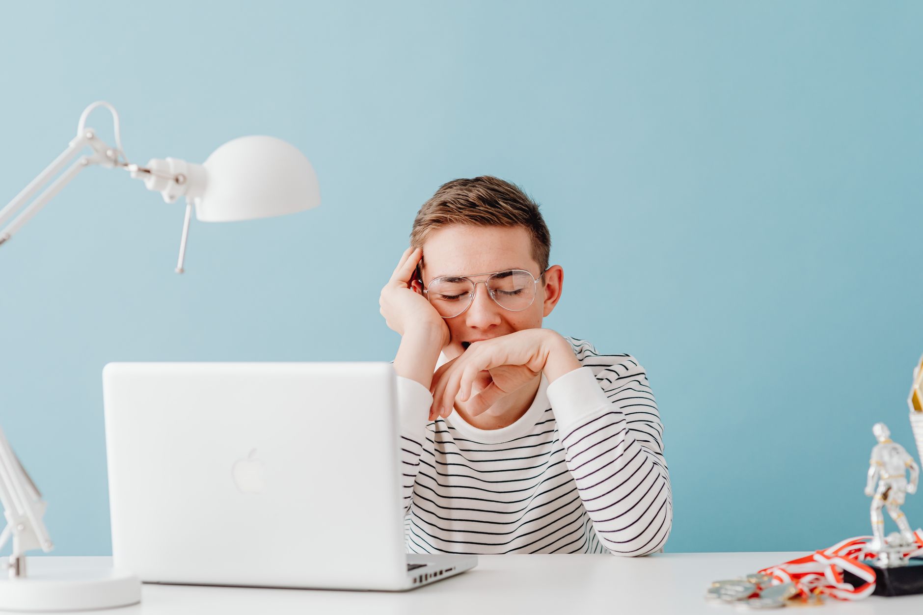 Young man in glasses yawning at desk with laptop and lamp, looking tired.