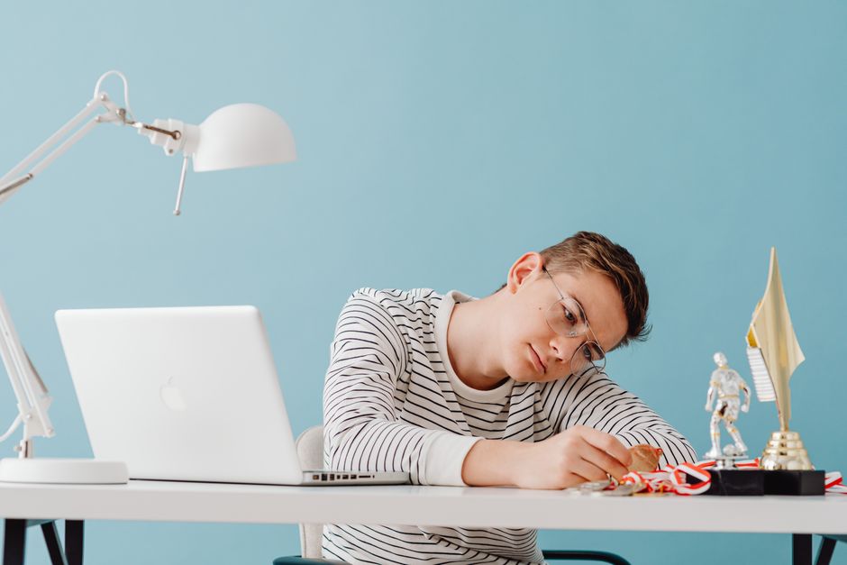 Teenager with eyeglasses contemplating medals at desk with laptop and trophies. Teenager with eyeglasses contemplating medals at desk with laptop and trophies.