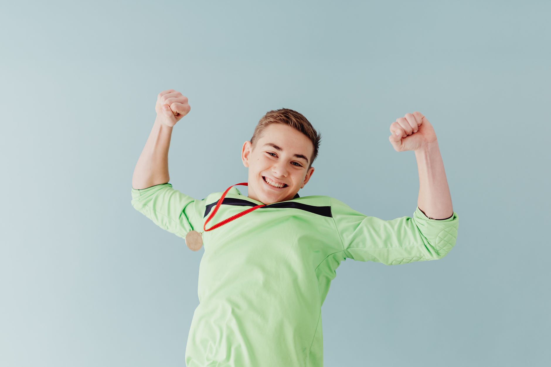 Excited teenage boy in green celebrates with a gold medal against a blue background.