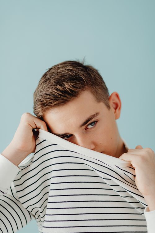 Free Teenage boy posing in studio with striped shirt, covering face lightheartedly. Stock Photo