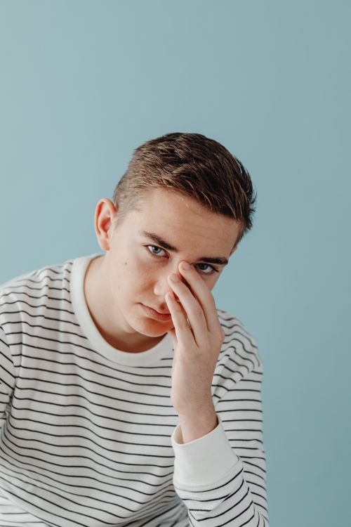 Free Teenage boy in striped shirt posing thoughtfully against a blue backdrop. Stock Photo