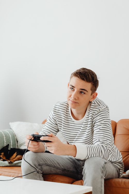 Free Teenage boy sitting on a sofa playing video games with a focused expression. Stock Photo