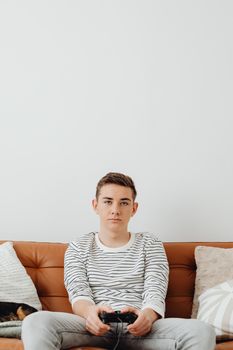 Teen boy focused on playing video games at home, seated on a modern leather sofa.