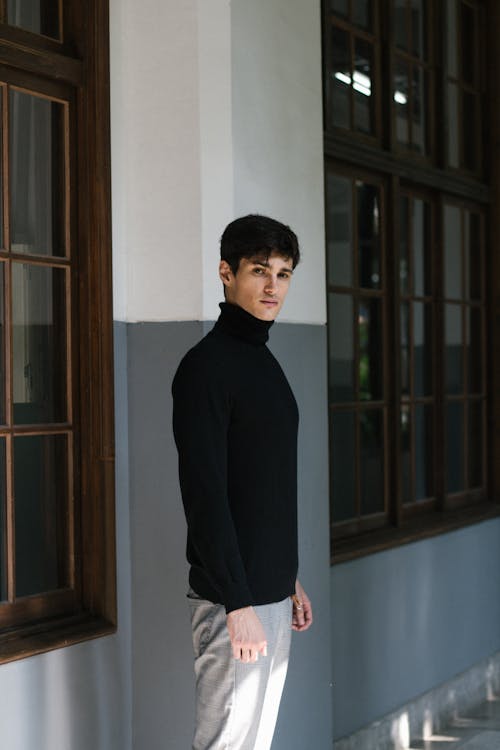 Free Side view of young male in black sweater looking at camera near windows of house in daytime Stock Photo
