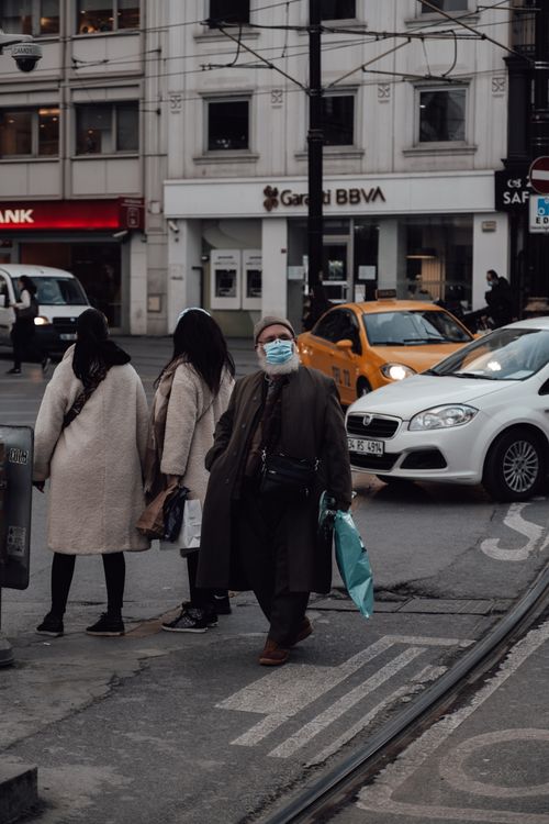 Free Old man in mask walking on street near pedestrians and automobiles driving on asphalt road during coronavirus pandemic Stock Photo