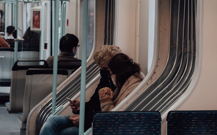 Side view of friends in protective masks sharing mobile phone while riding in metro during pandemic