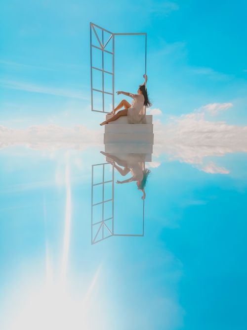Free A woman in a white dress relaxes by an open window with a surreal reflection under a blue sky. Stock Photo