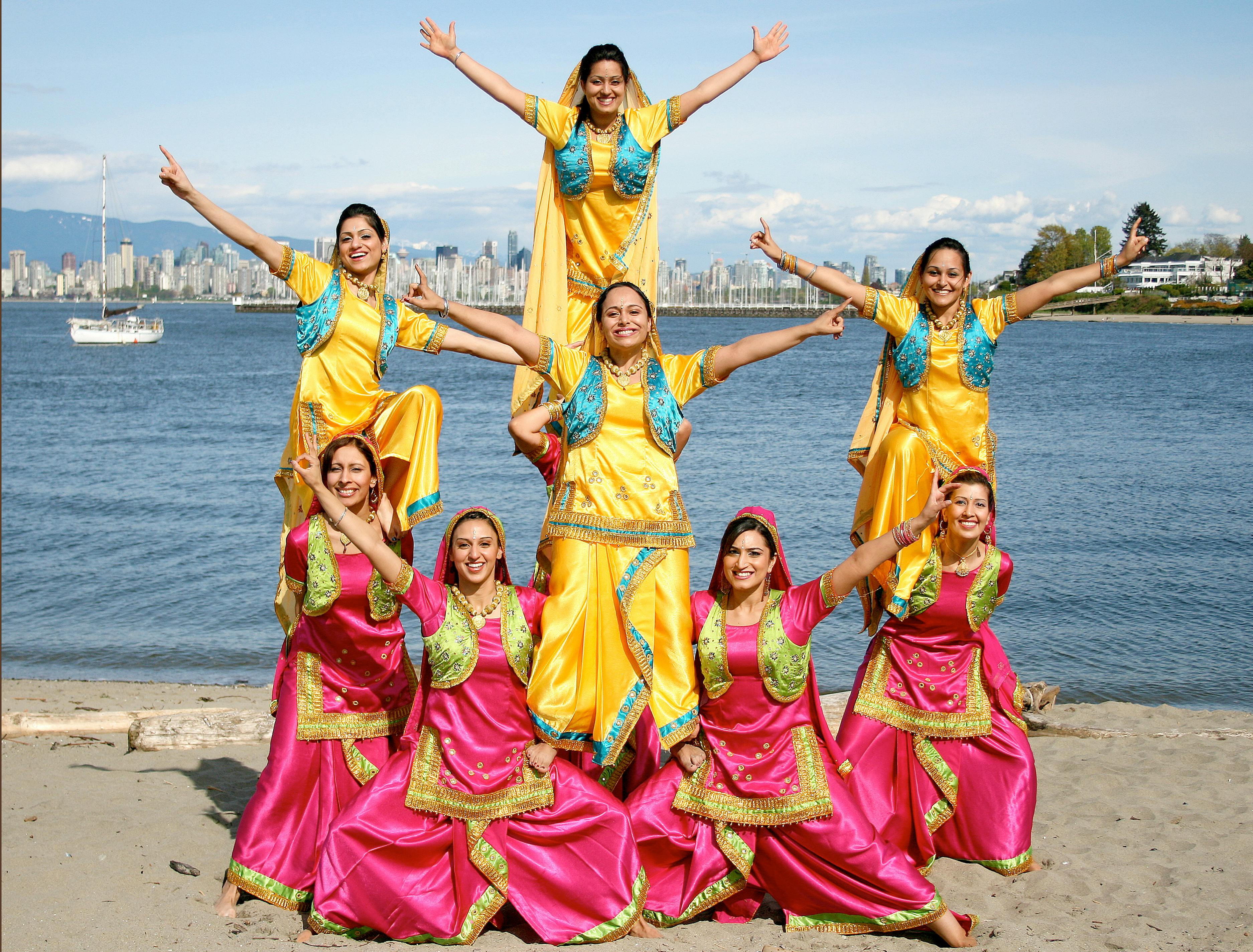 A joyful group of women performs a traditional dance on a sunny beach with a cityscape backdrop.