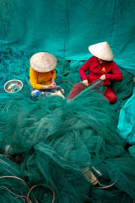 Authentic coastal community life in Quang Ninh, Vietnam, local fishers repairing nets by the sea, wooden boats nearby, weathered hands, salty atmosphere, sustainable livelihood storytelling, golden afternoon light, highly detailed